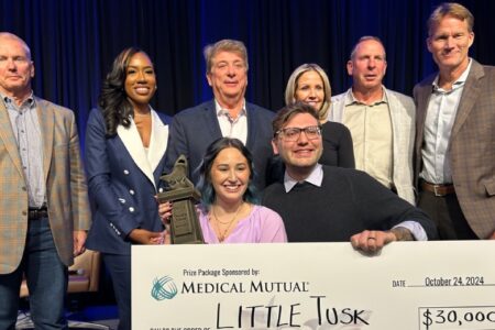 Lauran and Jordan Woolley display one of the cash prize awards their company, Little Tusk, won at Thursday’s Shark Tank competition. Behind them are judges, from left, Don Thomas, managing partner of Platz Realty;Ashley Vidale, co-owner of Sapphire Aesthetics and Wellness; Sam Boak, president of Boak & Sons Inc.; Stacy Howlett, CEO of Howlett Logistics; Bo Pelini, investor and former head football coach at the University of Nebraska and Youngstown State University; and Rob Cochran, chairman of #1 Cochran.